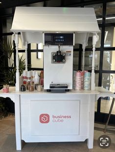 a small white stand with drinks on it in front of a window and plants behind the counter