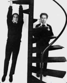 two men standing on top of a spiral staircase next to each other, one holding up the handrail