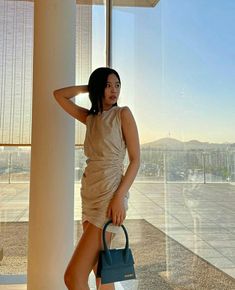 a woman standing next to a window holding a handbag and posing for the camera