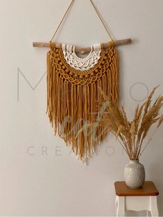 a white vase sitting on top of a wooden table next to a wall hanging with tassels