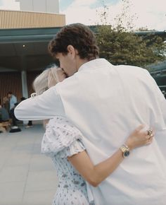 a man and woman embracing each other in front of a building with people sitting on benches