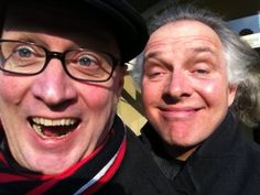 two men are smiling for the camera with one man wearing a hat and scarf around his neck