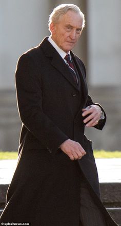 an older man in a long black coat and tie walking down the street with his hand on his hip