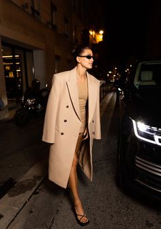 a woman is walking down the street at night wearing a tan coat and matching heels