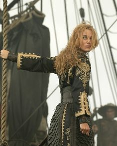 a woman dressed in black and gold standing on top of a boat with her arms outstretched