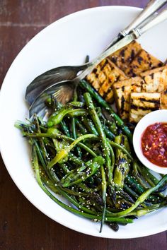 a white plate topped with green beans and grilled tofu next to a small bowl of sauce