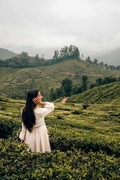 a woman standing in the middle of a lush green field with mountains behind her and clouds overhead