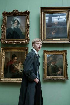 a young man standing in front of paintings on the wall and wearing a long coat