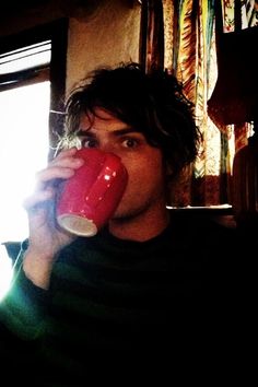 a man drinking from a red cup in front of a window with sunlight streaming through it