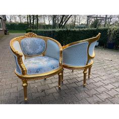 two blue and gold chairs sitting next to each other on a brick walkway with hedges in the background