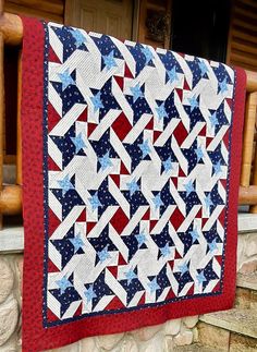 a red, white and blue quilt hanging from a wooden porch rail next to a stone wall