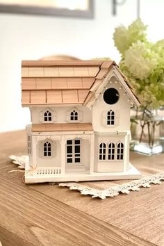 a small white house sitting on top of a wooden table next to a vase with flowers