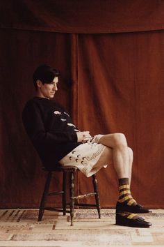 a man sitting on top of a wooden chair in front of a brown wall wearing socks