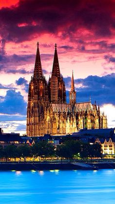 the cathedral is lit up at night with bright clouds in the sky above it and water below