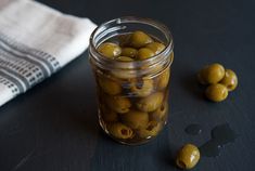 a glass jar filled with green olives on top of a black table next to a white towel