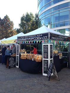 an outdoor market with food on display and people standing around it in front of a large glass building