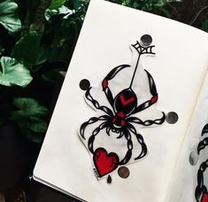 an open book with a spider and heart decorations on the pages, sitting next to some plants