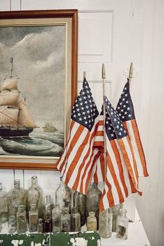 two american flags are next to an old painting and bottles on a shelf in a room