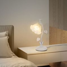 a white flower lamp sitting on top of a bed next to a night stand with pillows