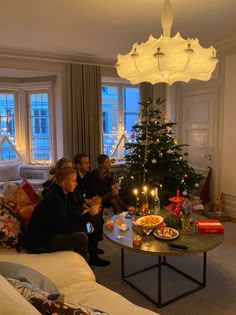 three people sitting around a table with food and candles in front of the christmas tree