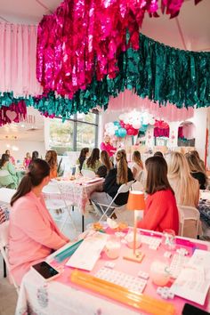 a group of people sitting at tables in a room with pink and green decorations hanging from the ceiling