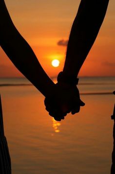 two people holding hands with the sun setting in the distance behind them, over water