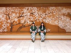 two people sitting on a bench in front of a wall with flowers and trees painted on it