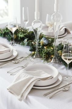 the table is set for christmas dinner with white plates, silverware and greenery