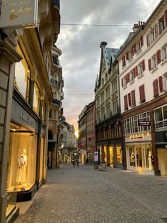 a cobblestone street lined with buildings and shops at dusk in the middle of town