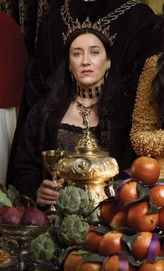 two women dressed in medieval clothing stand behind a table with fruit and vegetables on it