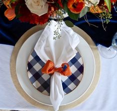 a white plate topped with an orange and blue napkin next to a vase filled with flowers