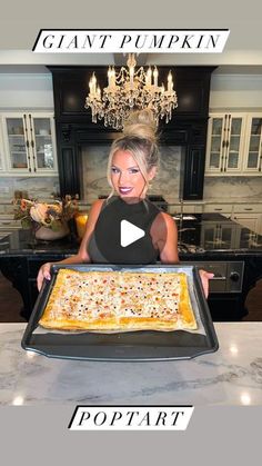 a woman holding up a large pizza on top of a pan in front of a chandelier