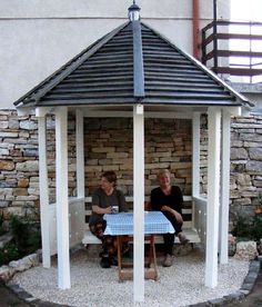 two people sitting at a picnic table under a gazebo in front of a stone wall