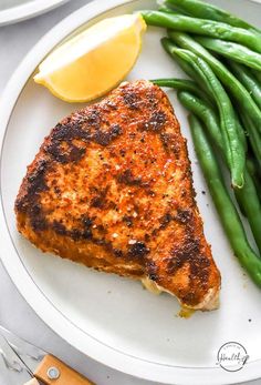 a white plate topped with green beans and a piece of meat next to a lemon wedge