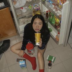 a woman sitting on the floor drinking from a cup in front of an open refrigerator