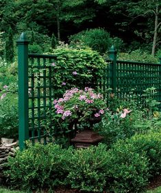 a green fence surrounded by shrubbery and pink flowers in the foreground is a potted plant with purple flowers on it