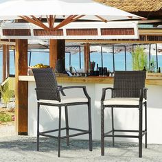 an outdoor bar with four chairs and two umbrellas over it, next to the ocean