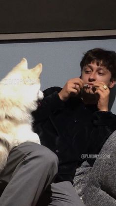 a man sitting on a couch next to a white dog eating something out of his mouth