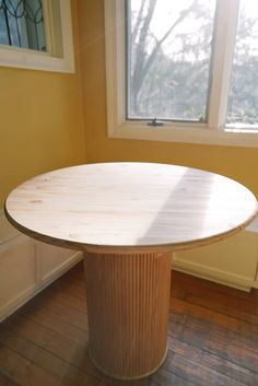 a round wooden table sitting on top of a hard wood floor next to a window