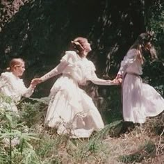 three women in white dresses holding hands and walking through the woods