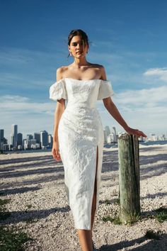 a woman in a white dress standing next to a wooden post on the beach with buildings in the background