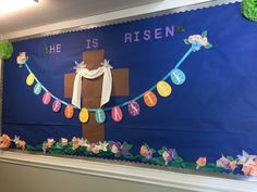 a bulletin board decorated with paper flowers and a cross in the center that says he is risen