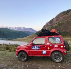 a small red car parked on the side of a mountain with mountains in the background