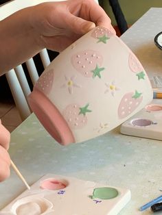 a person holding a cup on top of a table next to paints and pencils