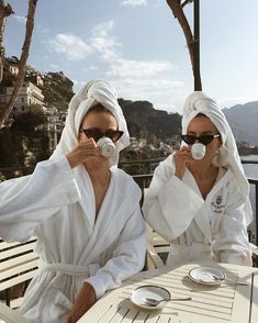 two women in white robes sitting at a table drinking from cups and saucers with their faces covered by towels