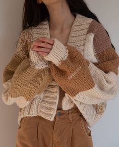 a woman standing in front of a white wall wearing a sweater and brown pants with her hands on her chest