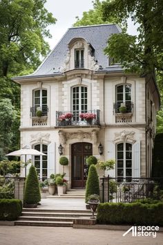 a large white house with lots of windows and plants on the front porch, surrounded by trees