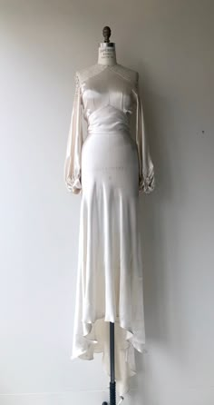 a white dress on a mannequin stand in front of a wall with an open back and long sleeves