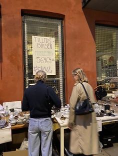 two people standing in front of a store with lots of items on the tables outside