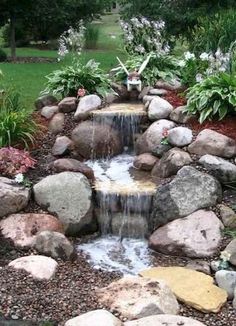 a small waterfall in the middle of a garden with rocks and flowers around it, surrounded by greenery